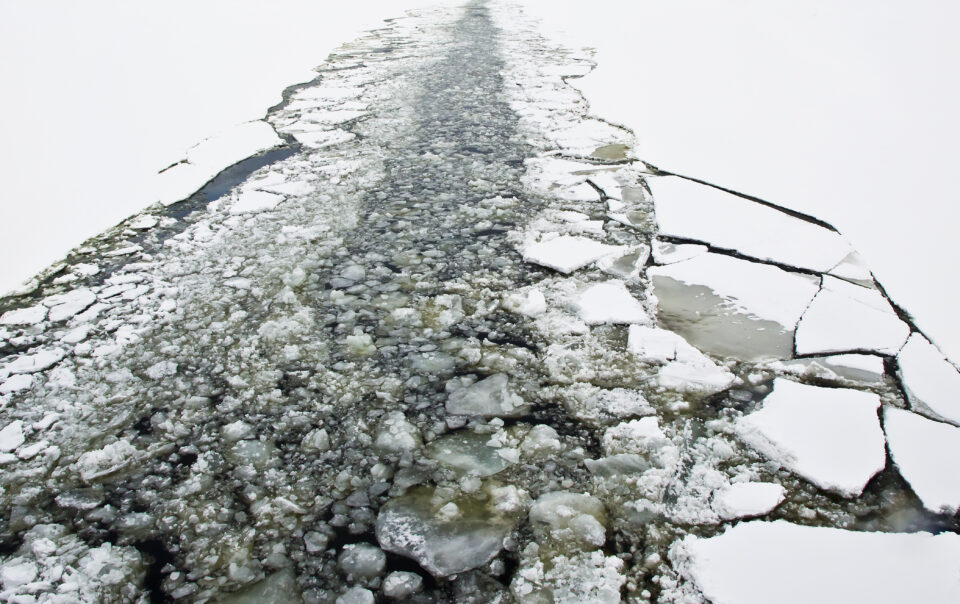 Eine aufgebrochene Schneedecke auf einem zugefrorenen Gewässer zeigt eine schmale, unregelmäßige Fahrrinne mit zerbrochenem und aufgeschobenem Eis.